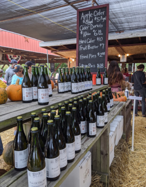 A market stall displaying bottles of apple cider, cider donuts, and pies, with pumpkins in the background.