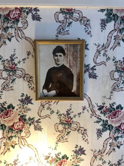 A vintage portrait of a woman in a black dress, framed and hung on floral wallpaper with ribbons and roses.