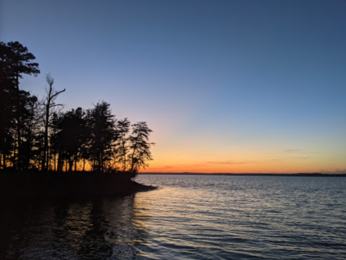 Silhouette of trees along a calm lake at sunset, with a gradient sky transitioning from orange to deep blue.