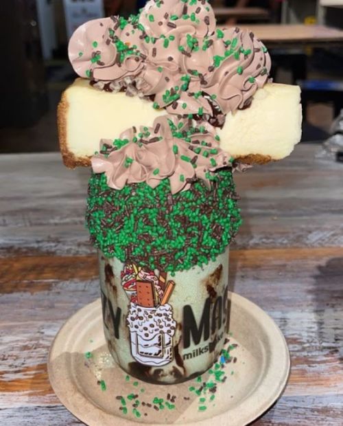 A tall milkshake topped with chocolate frosting, sprinkles, and a slice of cake, served in a branded glass.