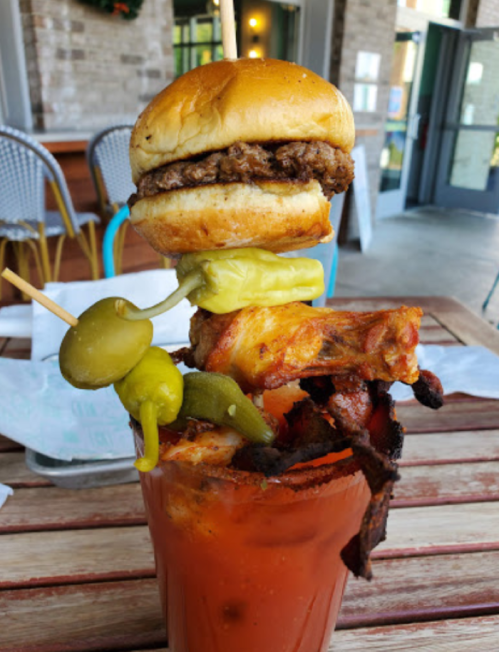 A towering Bloody Mary garnished with a mini burger, pickles, and various meats on a skewer, served in a glass.