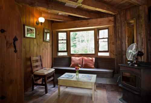 Cozy wooden interior with a bench, chair, table, and a small stove, surrounded by windows and natural light.