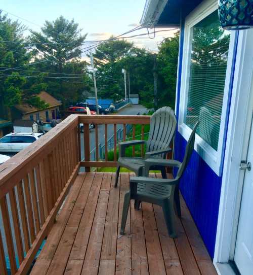 A wooden deck with a single gray chair, surrounded by trees and distant buildings under a clear sky.