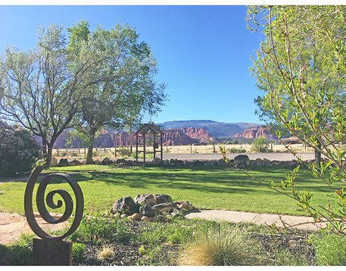 A scenic view of a grassy landscape with trees, a spiral sculpture, and distant red rock formations under a clear blue sky.