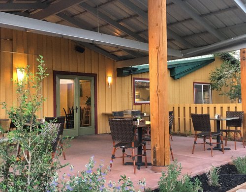 Outdoor dining area with wooden posts, tables, and chairs, surrounded by greenery and a rustic building.