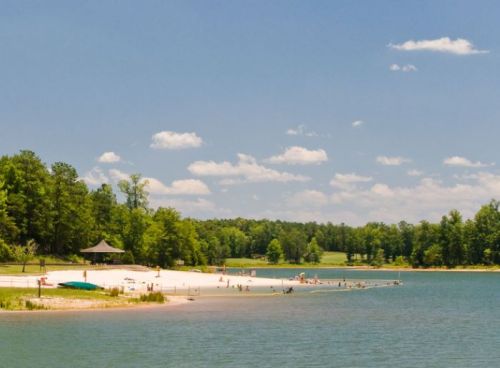 A serene lake scene with a sandy beach, green trees, and people enjoying the water under a clear blue sky.