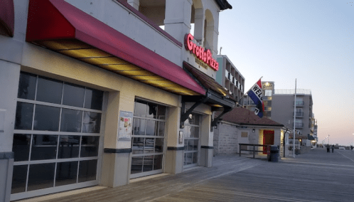 A boardwalk restaurant with a red awning, near the beach, at dusk. Flags and buildings are visible in the background.
