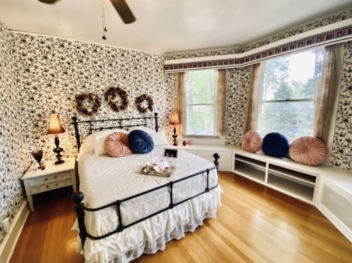 Cozy bedroom with floral wallpaper, a wrought iron bed, decorative pillows, and large windows letting in natural light.