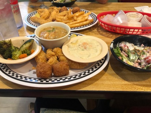 A table with two plates of food: fried shrimp, fries, salad, and sides of broccoli and cornbread. Drinks in the background.