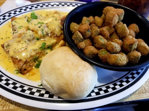 A plate featuring grilled fish with creamy sauce, a side of fried okra, and a soft roll on a checkered platter.