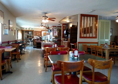 Cozy diner interior with wooden tables and chairs, ceiling fans, and a counter in the background.
