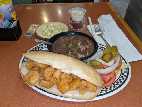 A plate featuring a shrimp po' boy sandwich, coleslaw, a bowl of beans, and pickles on the side.
