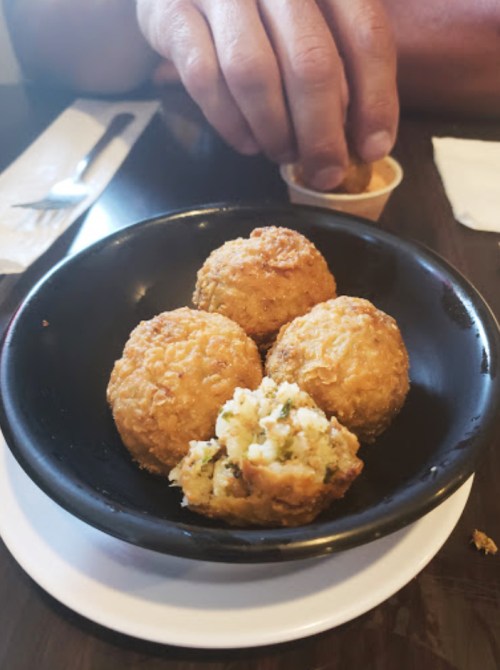 A black bowl with four golden-brown fried balls, one partially broken open, on a table with utensils in the background.