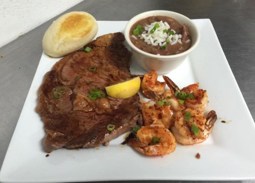 A plate featuring a grilled steak, shrimp, a side of beans, and a roll, garnished with green onions and lemon.