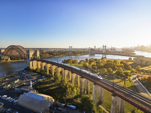 A train crosses a bridge over a river, with city skyline and autumn trees in the background under a clear sky.