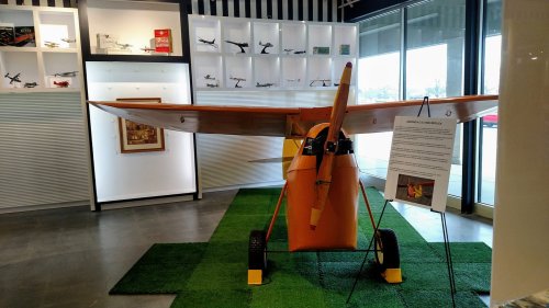 A vintage orange airplane model displayed indoors on green grass, with informational signage and framed images in the background.