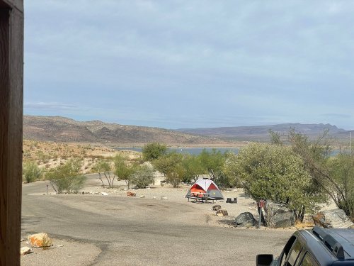 A scenic view of a campsite by a lake, featuring a tent and surrounding desert landscape under a cloudy sky.