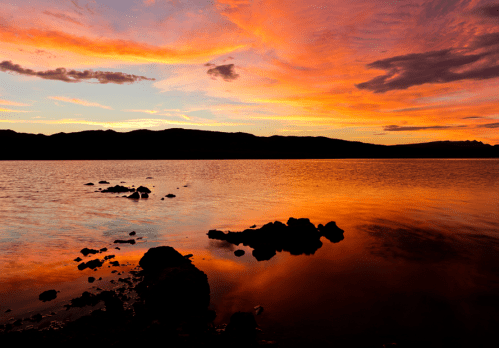 A vibrant sunset over a calm lake, with colorful clouds reflecting on the water and rocky shoreline in the foreground.