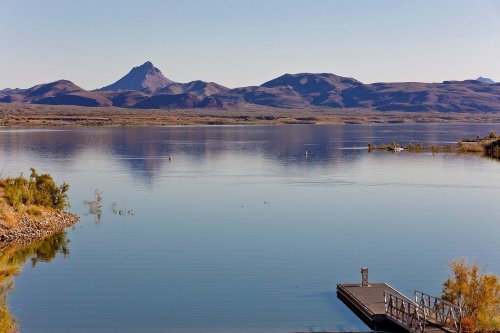 A serene lake reflects mountains under a clear sky, with a small dock in the foreground.