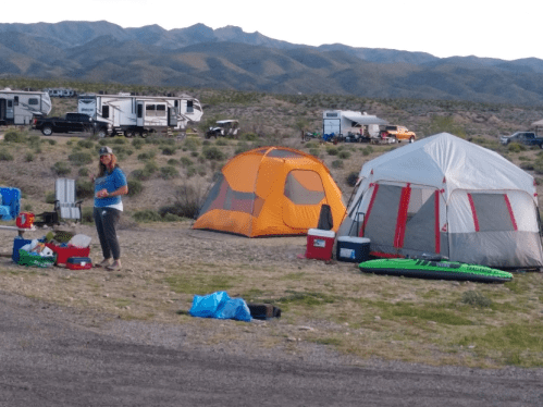 A campsite with two tents, a person standing nearby, and various camping gear scattered around in a desert landscape.