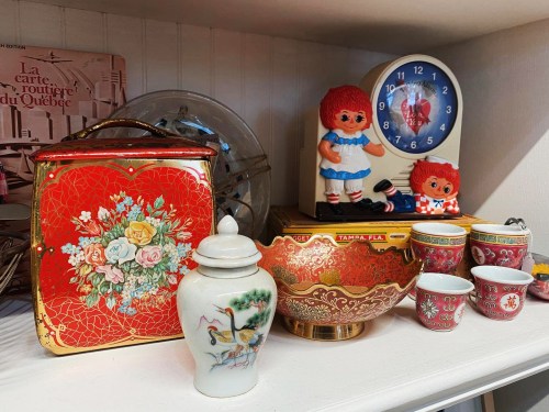 A shelf displaying vintage items: a red floral tin, porcelain vase, decorative bowl, and dolls with red hair.