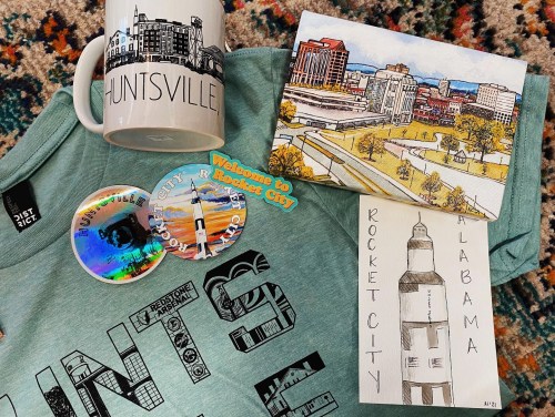 A collection of Huntsville-themed items: a mug, stickers, a T-shirt, and two illustrated postcards.