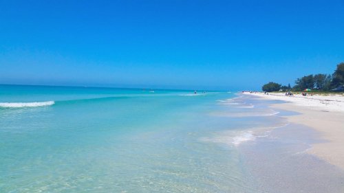 A serene beach with clear turquoise water, gentle waves, and a bright blue sky, lined with trees in the distance.