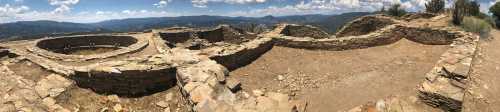 Panoramic view of ancient stone ruins on a hillside, surrounded by mountains and blue sky with scattered clouds.