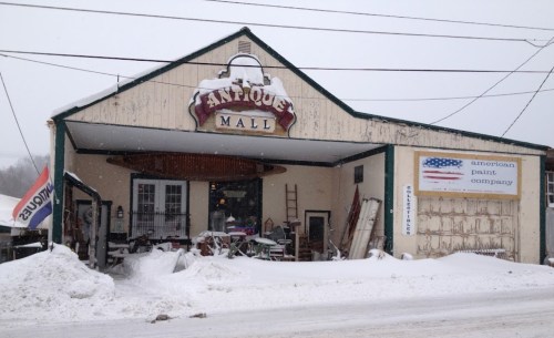 A snowy scene of an antique mall building with a sign, surrounded by piles of snow and a cloudy sky.