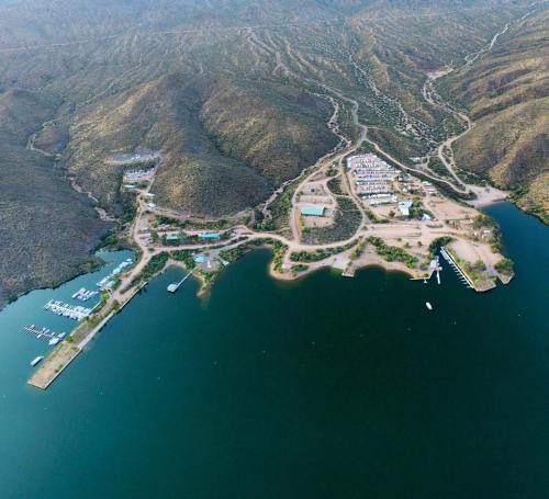 Aerial view of a marina and surrounding hills, featuring boats docked along the shoreline and a small settlement nearby.