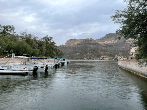 A calm river scene with boats docked along the shore, surrounded by trees and rocky hills under a cloudy sky.
