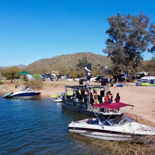 A scenic lakeside view with boats, people enjoying the outdoors, and tents under trees in a sunny environment.