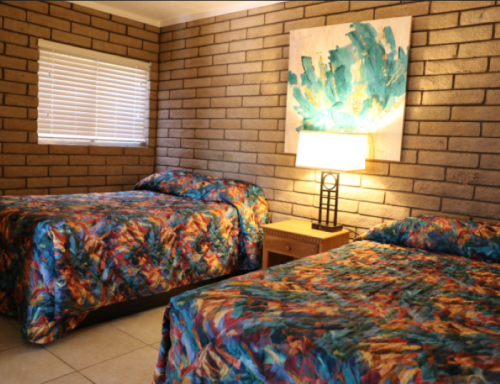 Two beds with colorful patterned bedspreads in a room with brick walls and a lamp beside a piece of abstract art.