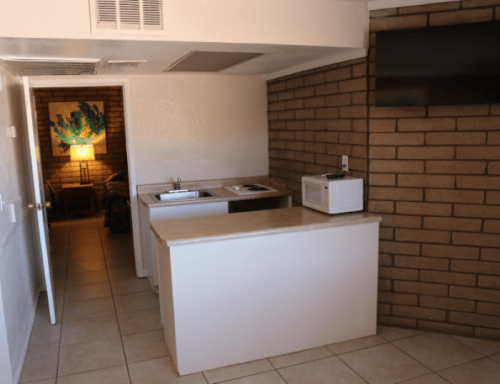 A small kitchen area with a countertop, sink, microwave, and a TV on the wall, featuring brick walls and tiled flooring.