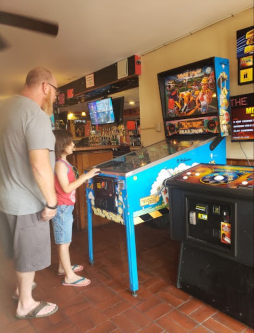A man and a girl play a pinball machine in a casual bar setting with screens in the background.
