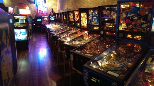 A row of vintage pinball machines in a dimly lit arcade, showcasing colorful designs and illuminated playfields.