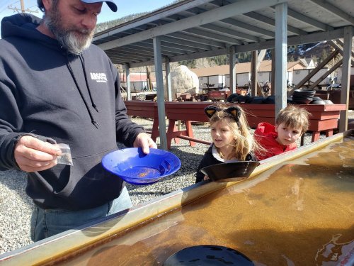 A man shows two children how to pan for gold at an outdoor mining site.