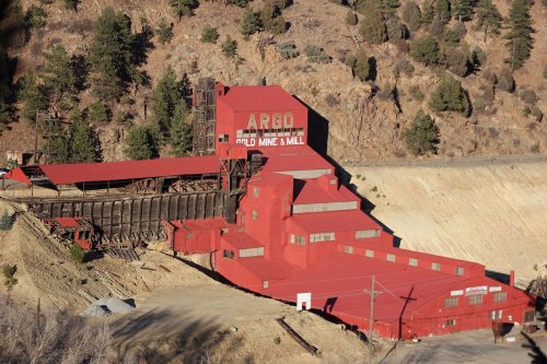 Aerial view of the Argo Gold Mine and Mill, a large red industrial building surrounded by trees and mountains.