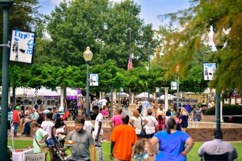A lively outdoor event with crowds, trees, and banners, set in a park with booths and activities.