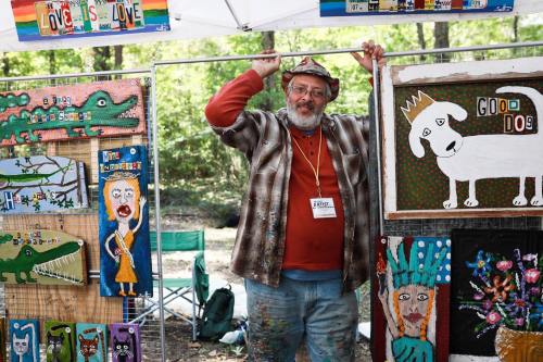 A smiling artist stands under a tent, showcasing colorful, whimsical artwork on wooden boards.