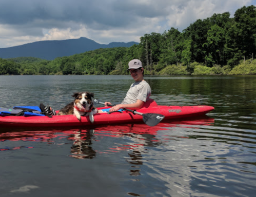 A person and a dog sit in a red kayak on a calm lake, surrounded by green trees and mountains under a cloudy sky.