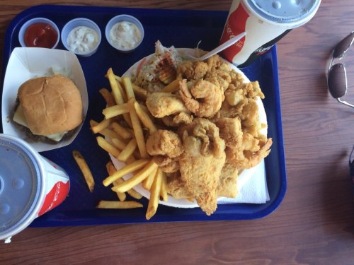 A tray with fried chicken, fries, a sandwich, and dipping sauces, alongside drinks and sunglasses.