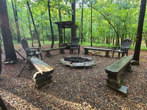 A serene outdoor fire pit area surrounded by trees, featuring stone benches and wooden chairs.