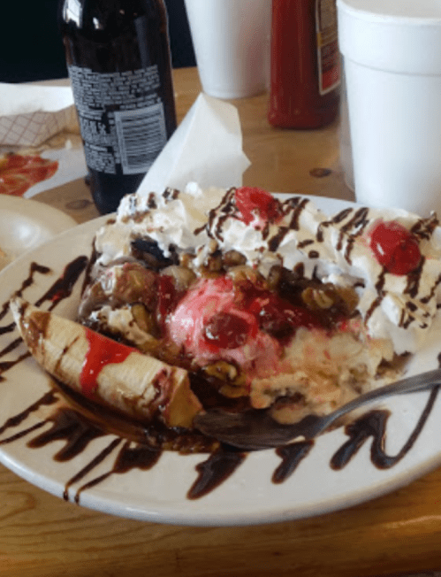 A dessert plate featuring banana split topped with whipped cream, cherries, nuts, and chocolate sauce.