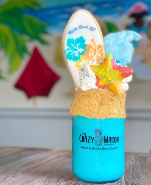 A colorful dessert in a jar, topped with whipped cream, a surfboard cookie, and star-shaped candy, with a beachy background.