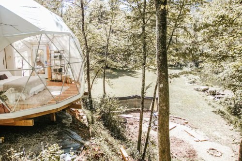 A geodesic dome cabin nestled among trees by a serene river, featuring a cozy interior and natural surroundings.