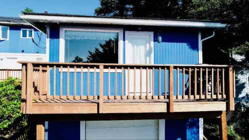 A blue house with a wooden deck and railing, featuring large windows and a white door.