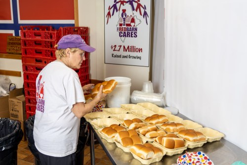 A woman arranges bread rolls on a table in a community kitchen, with a sign for Redbarn Cares in the background.
