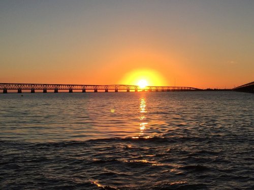 Sunset over a calm body of water, with a bridge silhouetted against the vibrant orange and yellow sky.