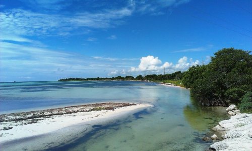 A serene coastal scene with calm waters, sandy shores, and lush greenery under a blue sky with scattered clouds.